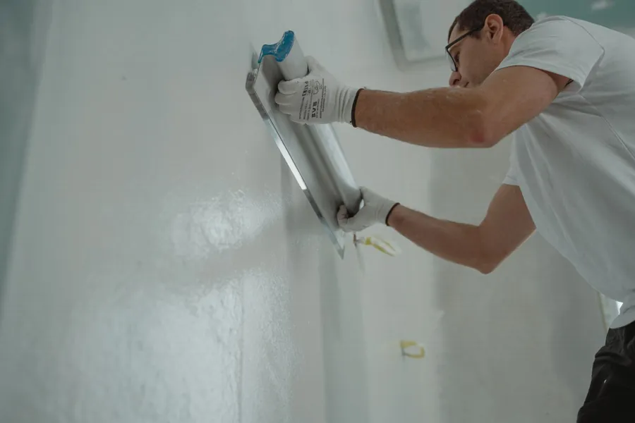 A plasterer working a wide trowel across a freshly skimmed wall, the making-good that sometimes has to happen before a flat roof flashing can be fitted