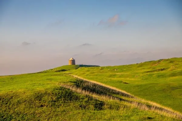 A lone stone tower on a green hilltop path, the kind of silhouette St Michael's tower cuts on Glastonbury Tor